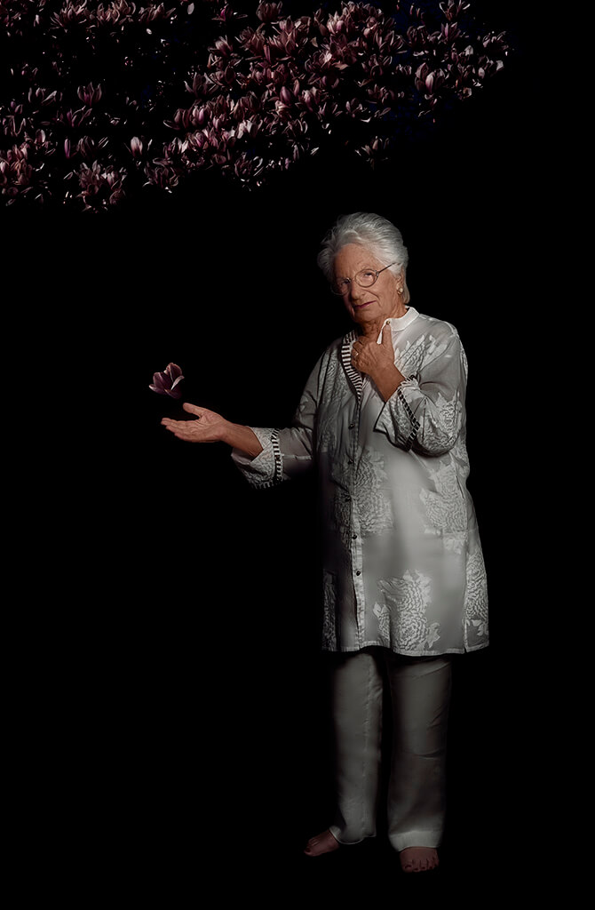 An older woman with white hair stands in a white tunic against a pitch black background. She holds out one hand and a glimmering purple leaf floats above it. Above her is a glimmering canopy of purple leaves.