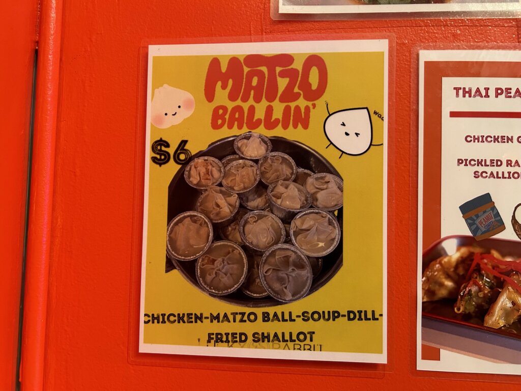 A poster with the words "Matzo Ballin'" at the top, above a pile of small aluminum shot glasses filled with dumplings covered in broth and garnished with fried shallot and dill. At the bottom, the words "chicken-matzo ball-souop-dill-fried shallot."