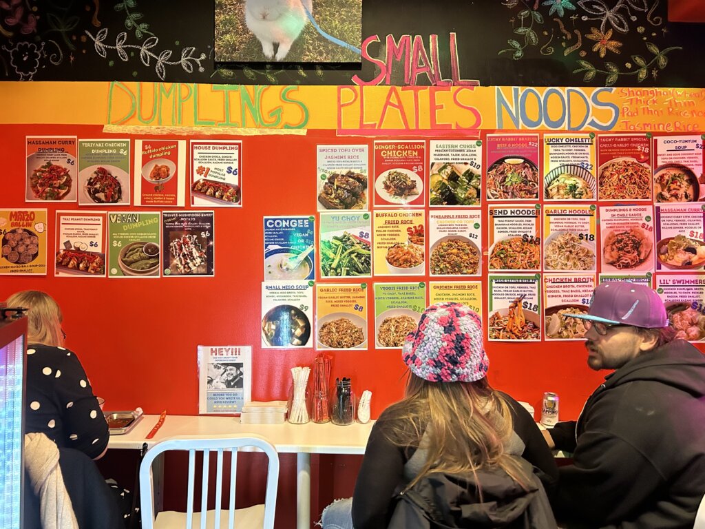 People sit around a counter against a red wall with the words, "dumplings," "small plates," and "noods" painted on top, and posters with the photos and names of dishes pasted all over the wall. Some examples: "Truffle mushroom sweet potato dumplings," "congee," and "garlic noodles."