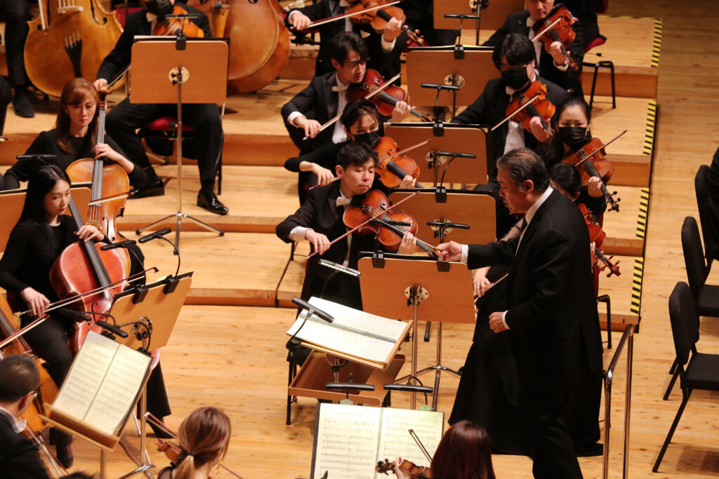 A man in a black suit conducts a row of violinists, with other performers at the edge of the frame.