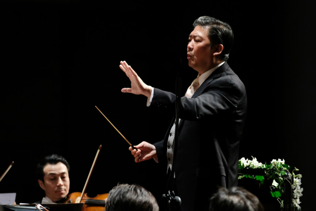 Man in suit holding left arm up, right arm holding conducting baton, against black background. Some people's heads poke into the background in the foreground, and in the background a man plays violin.