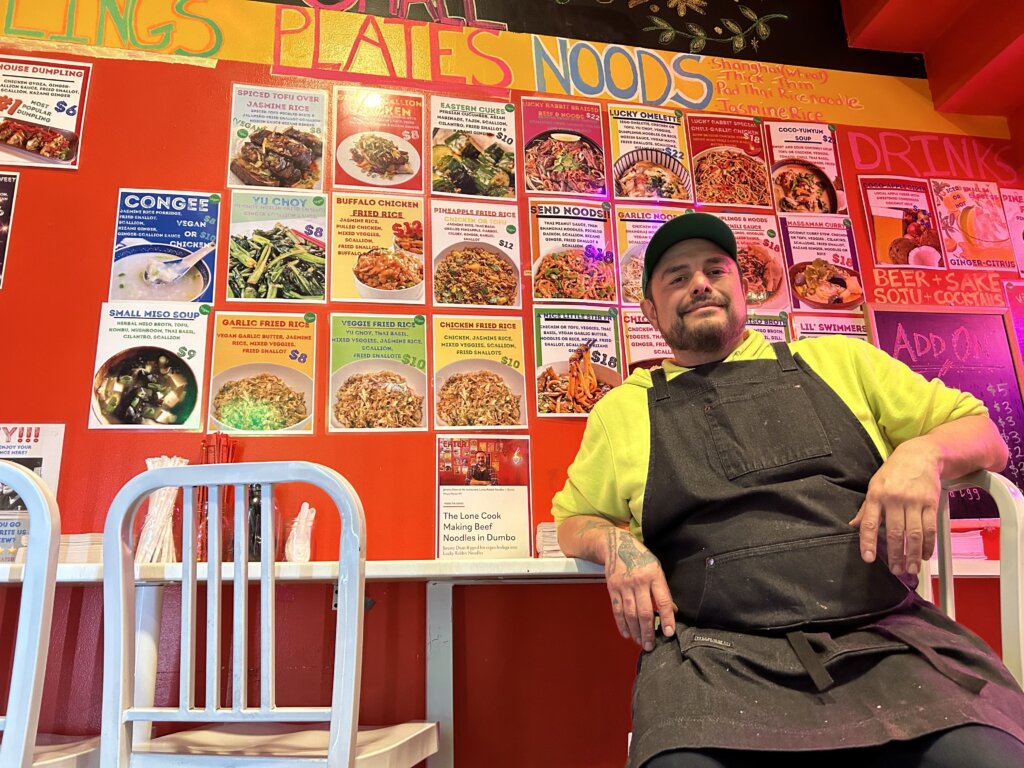 Man in denim overalls and neon yellow hoodie and green baseball cap leans back against a counter that abuts the red wall. on the wall, the words "Plates" and "Noods" are hand-painted at the top, above pieces of paper printed in different colors with names and photos of dishes, like "Yu Choy" greens, "Buffalo Chicken Fried Rice," and "Lucky Rabbit Braised Beef and Noods," a noodle soup dish.