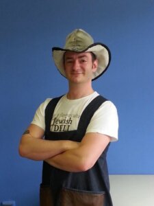 Man smiles with folded arms, wearing apron, white T-shirt, and a cowboy hat.