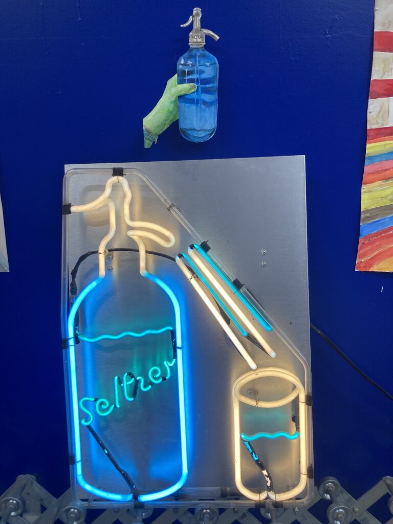 A neon sign with a seltzer bottle siphon that says "seltzer" and lines from the spout to a cup of a liquid.