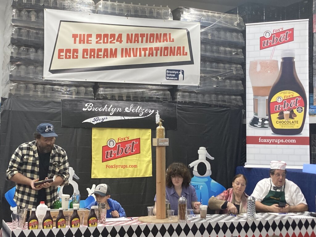 Several people stand behind at a long, horizontal table. Behind them, a sign that says "The 2024 National Egg Cream Invitational." In front of them, and another that says "brooklyn seltzer boys" and another that says "fox's u-bet; foxsyrups.com." There are several containers of syrup on the table. In the center, a large wooden pedestal with a golden siphon bottle at the top.