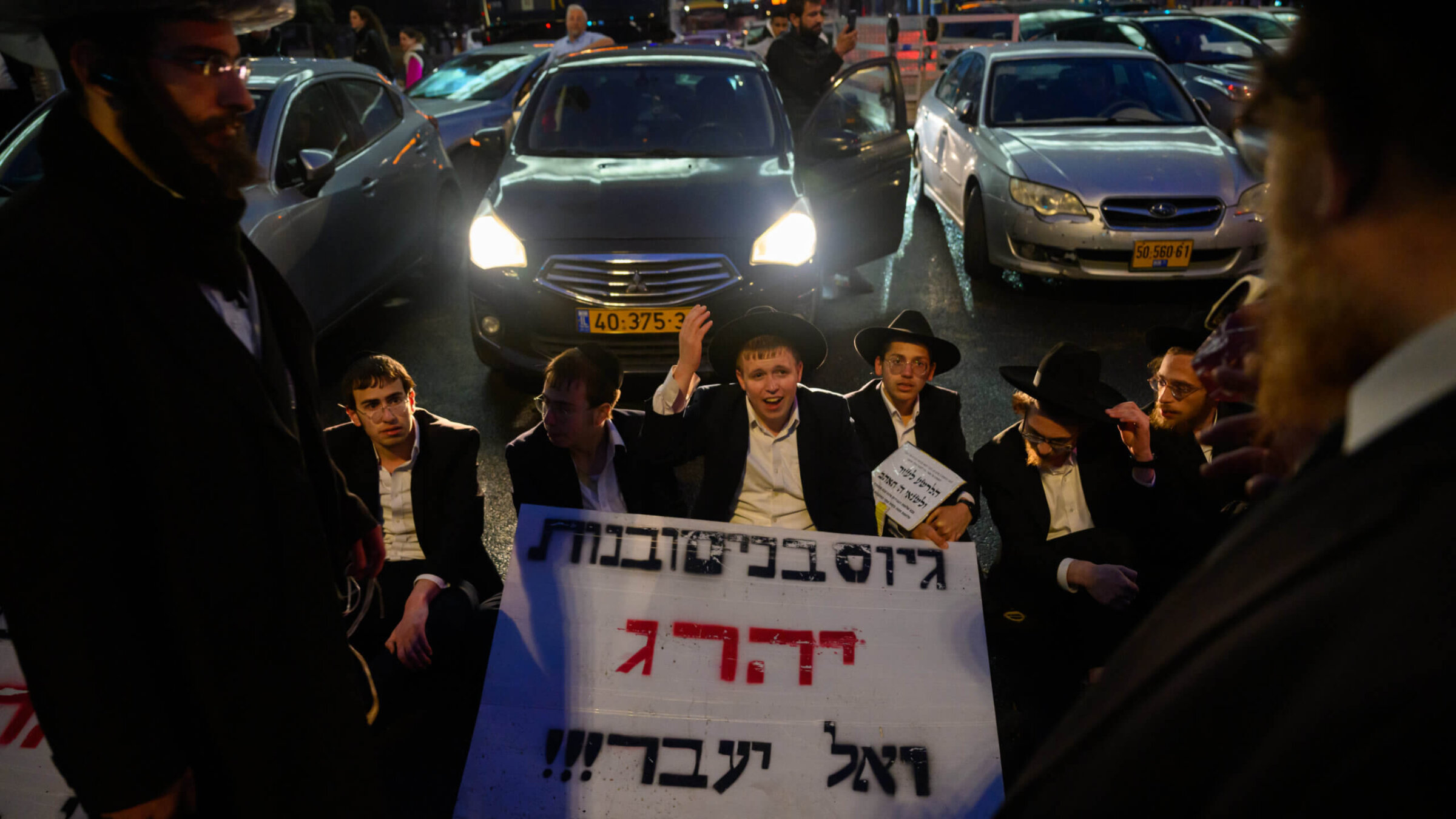 Haredi boys and men sit in front of traffic during a protest against the expiration of a law preventing them from being drafted into the IDF, March 18, 2024 in Jerusalem. 