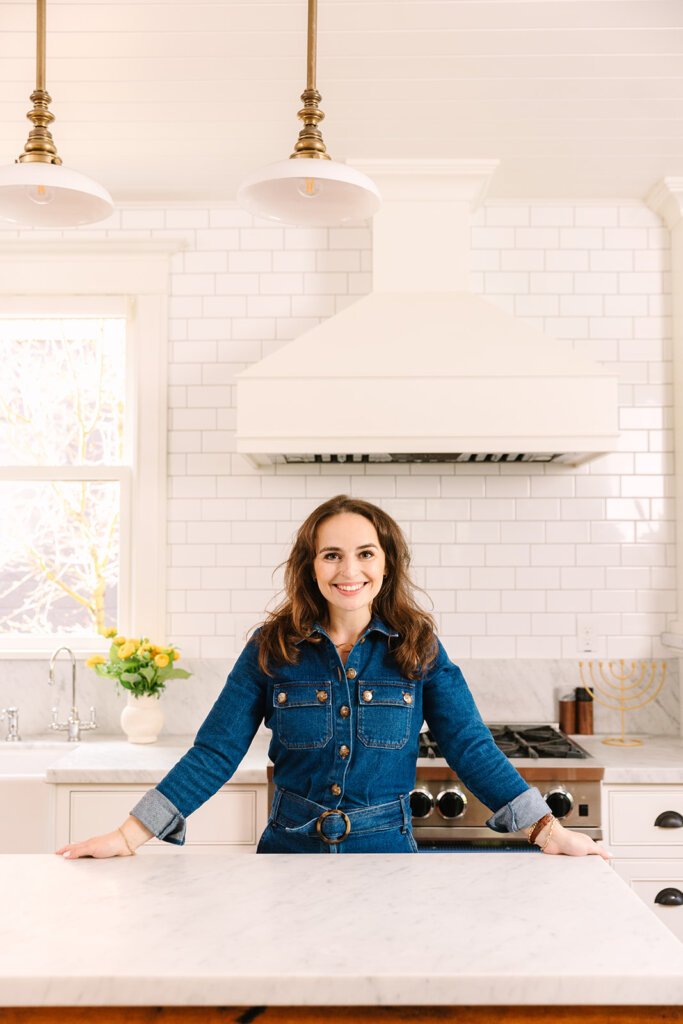 Woman in blue denim long sleeve shirt smiles while leaning on a white kitchen island with white tile kitchen behind her.