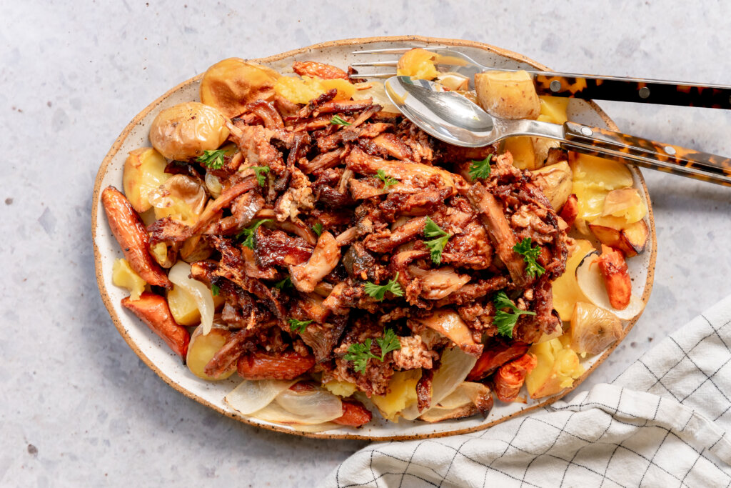 A top view of a plate of shredded, brownish-red brisket, with some noticeable chopped mushrooms, topped with curly leaf parsley and sitting on top of a bed of roast carrots and roast potatoes.