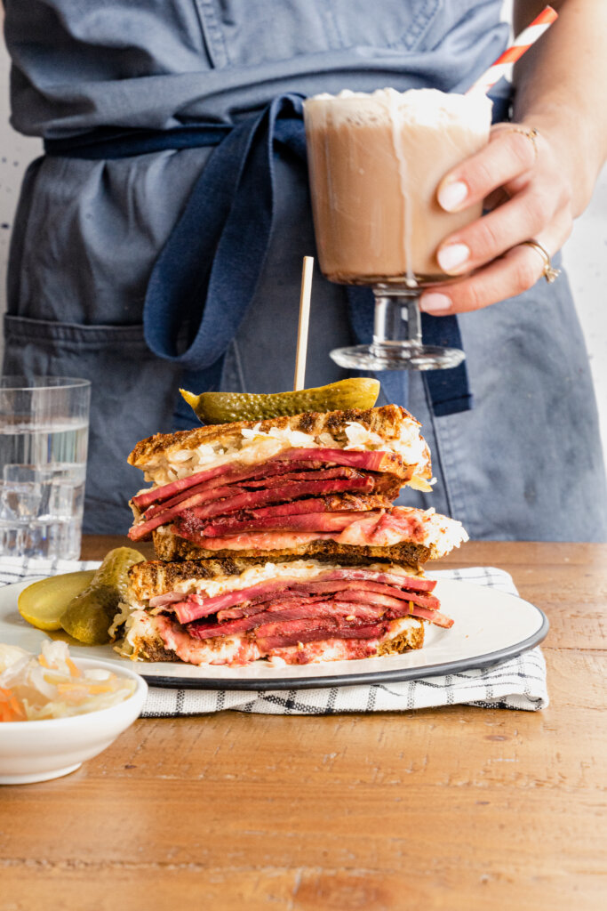 two sandwich halves, filled with pastrami, stacked one on top of the other, on a plate with pickles next to it. In the background, the torso of someone with an apron holding a glass with a brown, frothy liquid and a red and white straw.