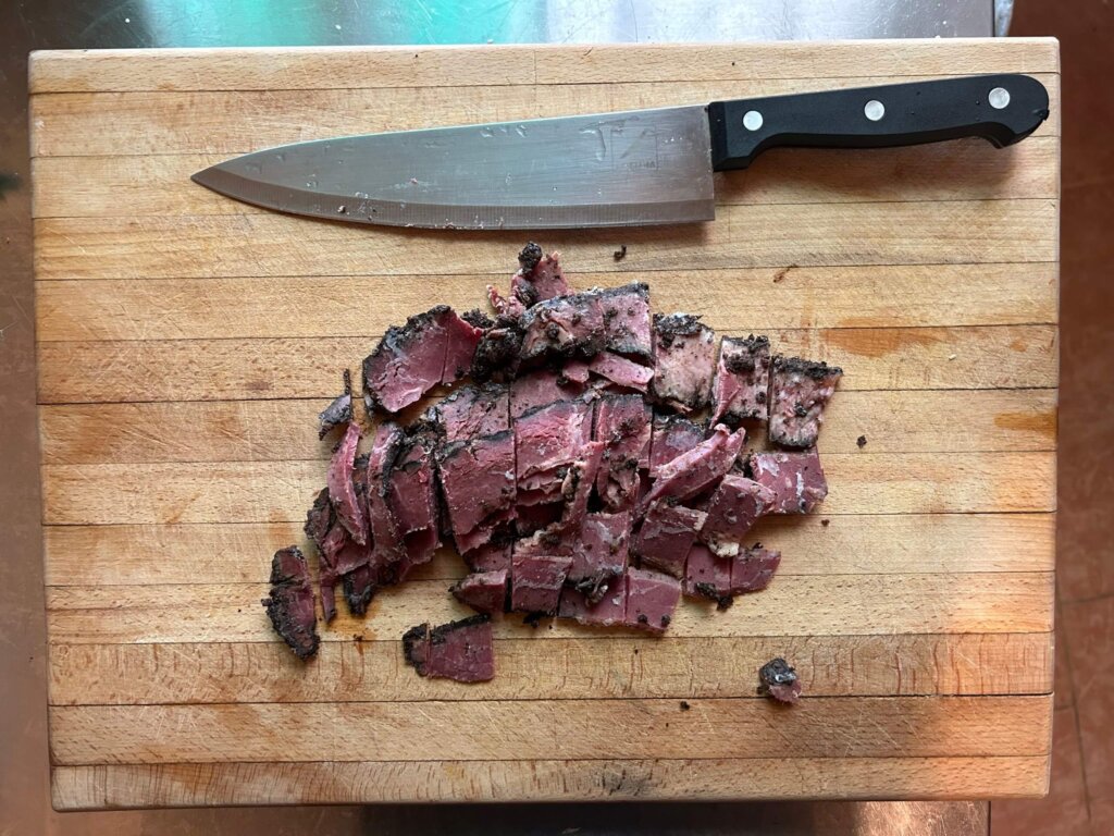 A wooden cutting board with chopped up pastrami and a large kitchen knife on it.