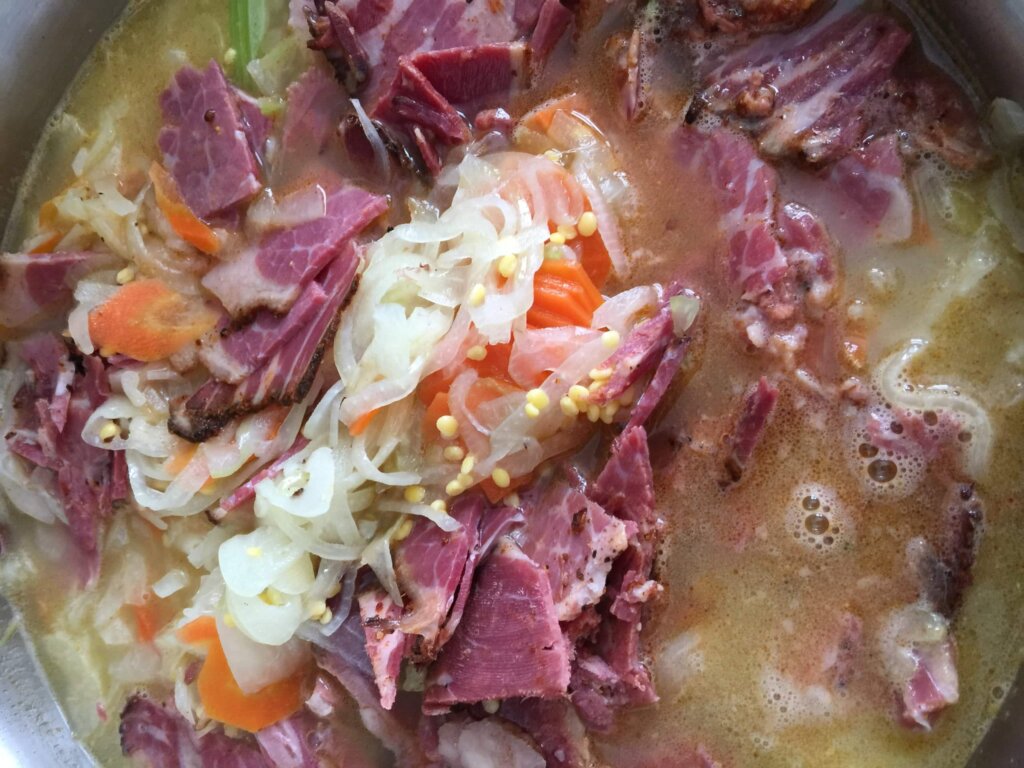 A close-up of a yellow broth topped with shaved red pastrami, sliced white shallots, and yellow mustard seeds.