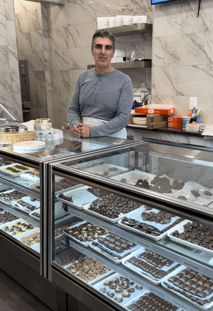 A man in a grey shirt stands behind a counter full of trays of chocolates and in front of a polished grey marble wall.