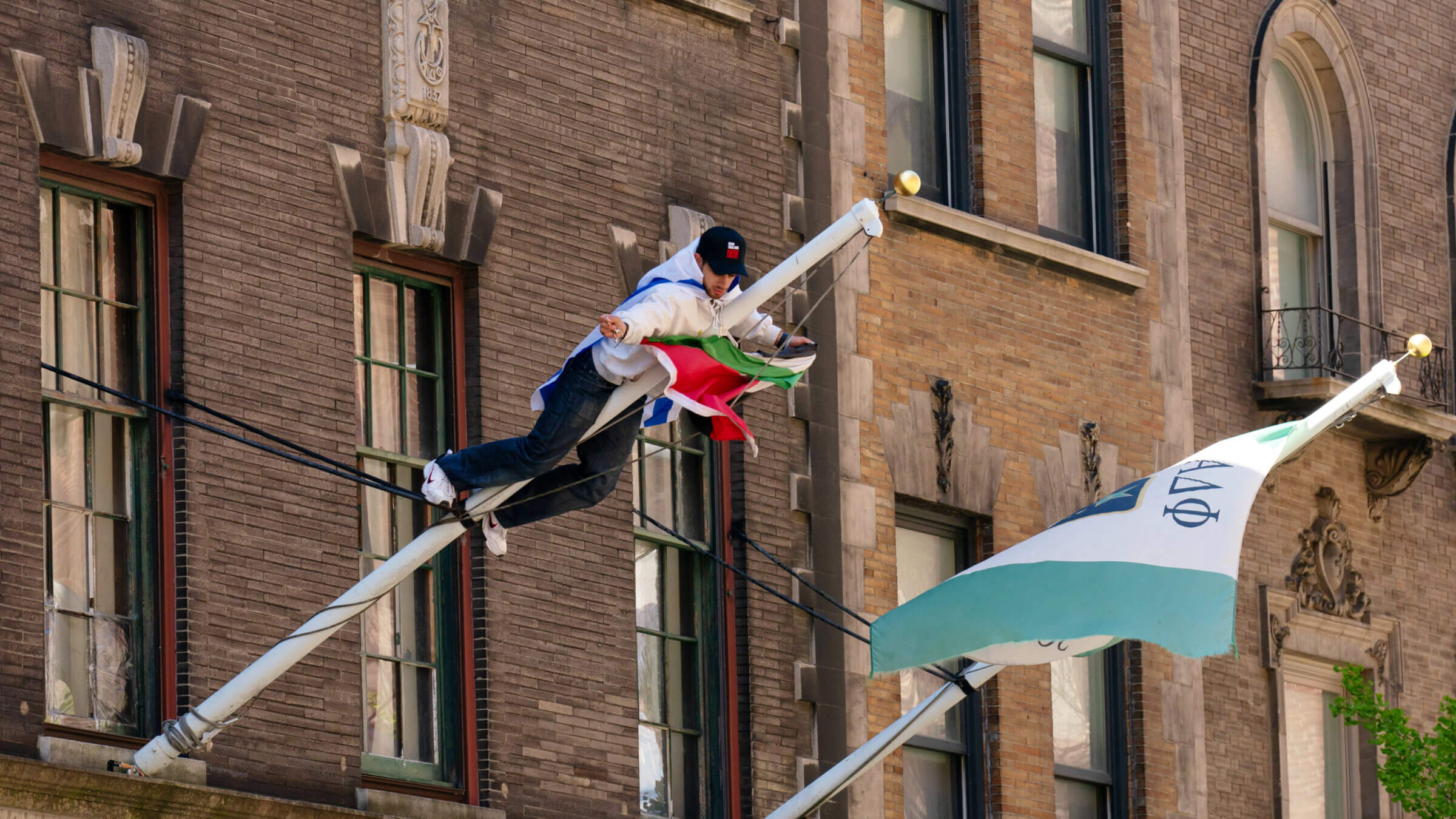 A man wearing an Israeli flag climbs a pole to remove a Palestinian flag from a Columbia University fraternity building on April 26, 2024 in New York City