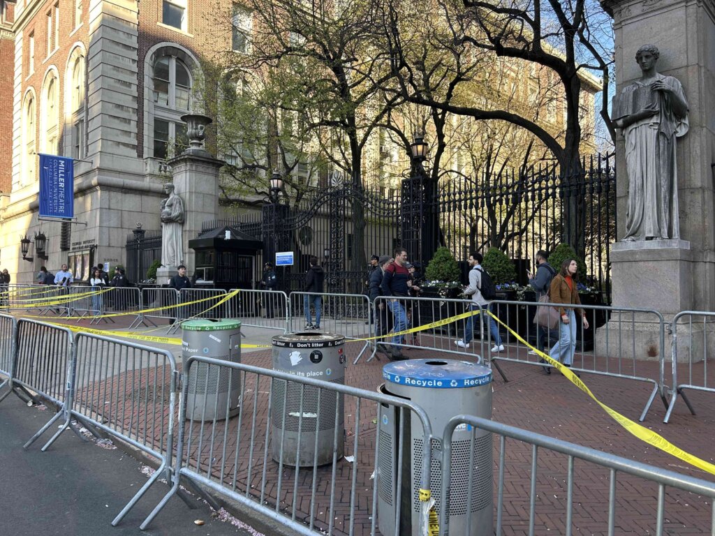 Police stand in front of iron gates, tall buildings on either side of them. In front of the gates, metal barriers arranged in a rectangle crisscrossed with yellow caution tape.