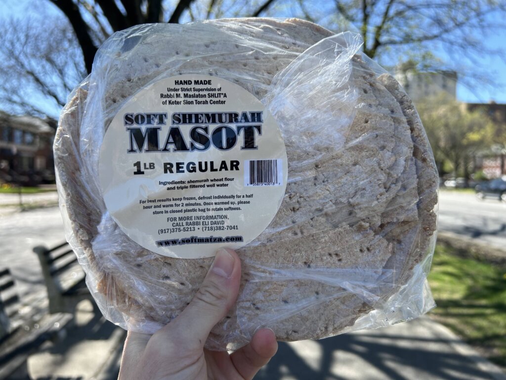 Hand outdoors holding a plastic package of round flatbreads with a label that says "Soft Shemurah Masot: 1 LB Regular" on it.