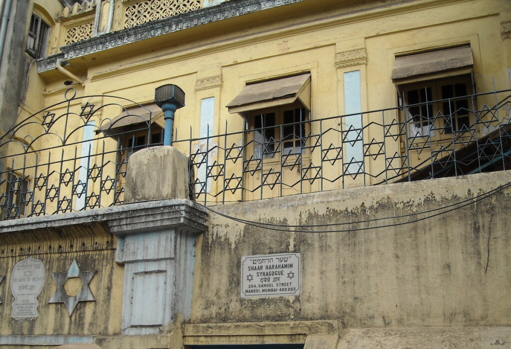 A yellow building with a gate made of stars of david in front of it and a plaque that reads, "Shaar Harahamim Synagogue," along with other words in Hebrew and Hindi, and 254 Samuel Street, Mandvi, Mumbai - 400 003