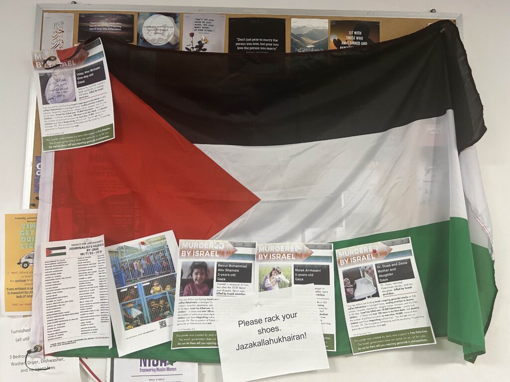 A Palestinian flag hanging on a wall and covered with posters that say "Murdered by Israel" with photos of people on them. Another page tacked on the flag reads "PLEASE RACK YOUR SHOES. JAZAKALLAHUKHAIRAN!"