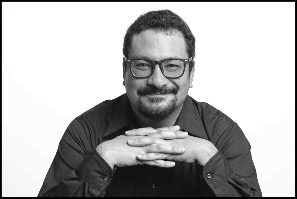 An Asian-Jewish man with a goatee and glasses smiles at the camera with his hands knit together below his chin. The photo is black-and-white.