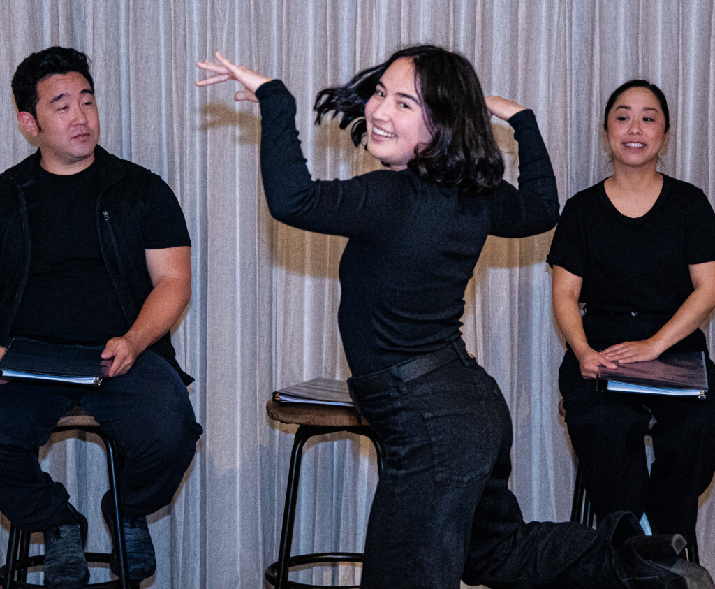 Two asian actors dressed in all black sit on stools in the background in front of a gray curtain. In the middle, one Asian actor dances and smiles.