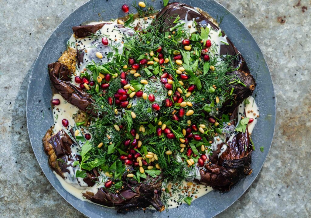 Blue plate with roasted eggplants covered in tahini, black sesame seeds, dill, parsley, pomegranate seeds and pine nuts.