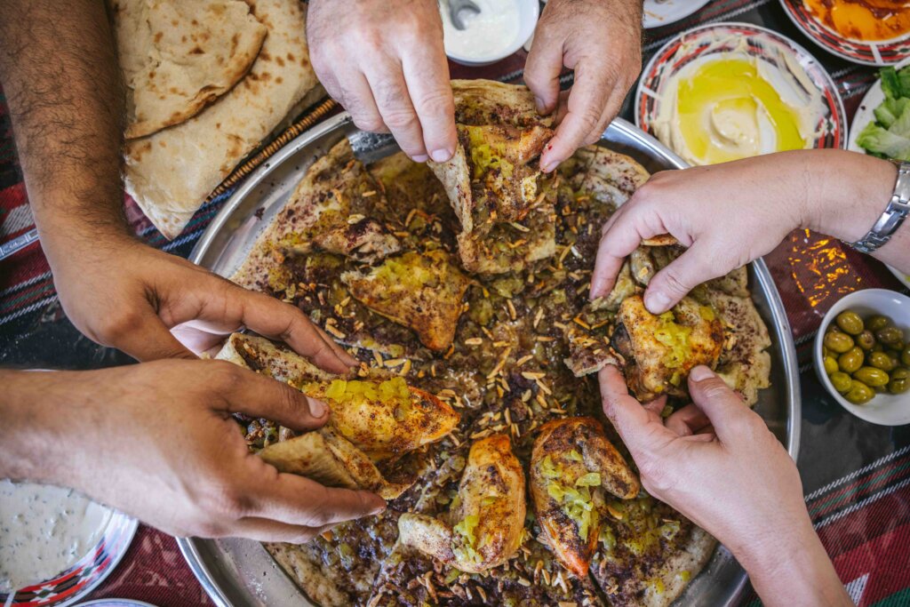 A circle of hands digging in to a dish with a large circular bread covered in spices and almonds and roast chicken.