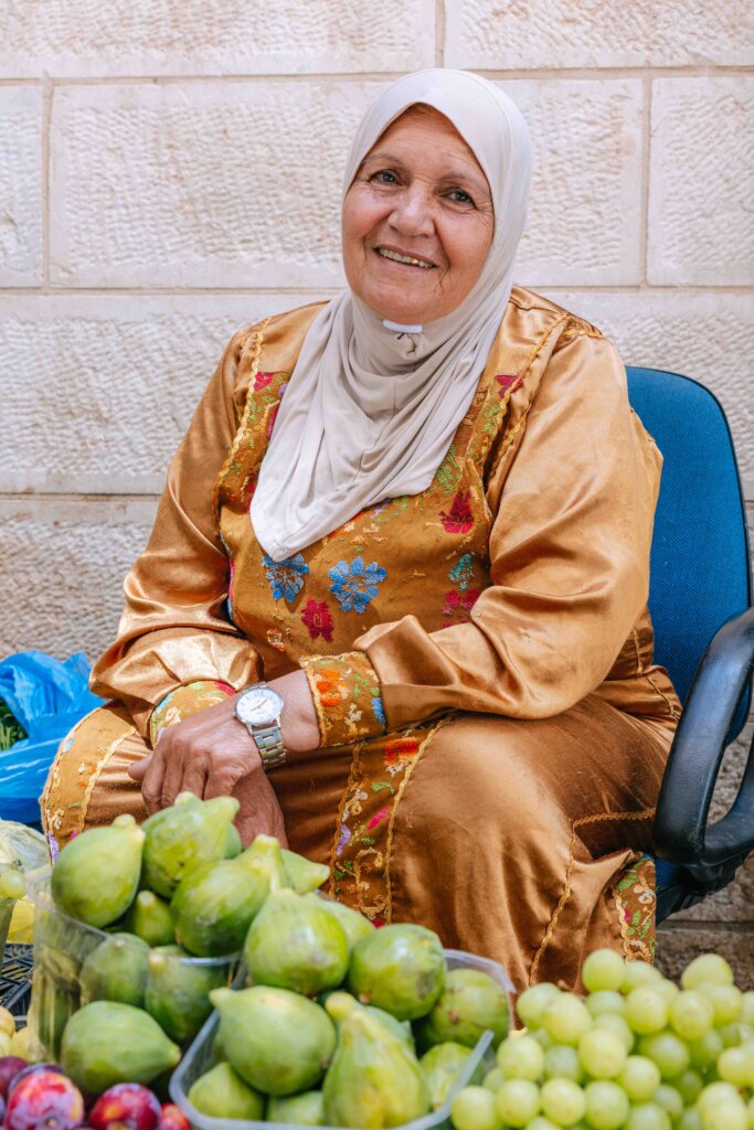 A middle aged woman wearing a hijab and an embroidered rust-colored robe sits on a chair and smiles with piles of grapes, nectarines, and another green pear-shaped fruit in front of her.