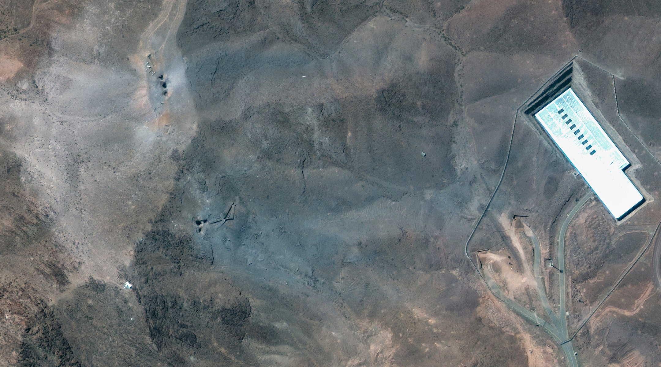 Maxar Satellite Imagery shows the ridge above the Fordow complex in Iran post-strike. Multiple impact craters and a wide grey-blue ash layer are visible across the ridge, suggesting direct hits to the overburden covering the underground facility. (Satellite image (c) 2025 Maxar Technologies)