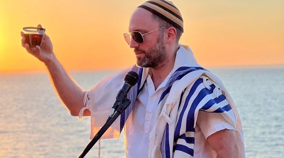 At sunset by the water, Rabbi Ari Yehuda Saks leads a blessing with wine in hand and tallit draped over the shoulders 