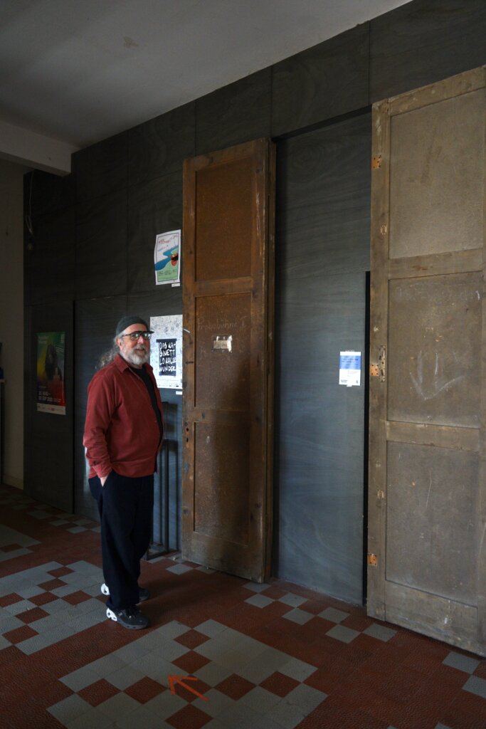 An old man in a red shirt stands next to a long door in a dark building
