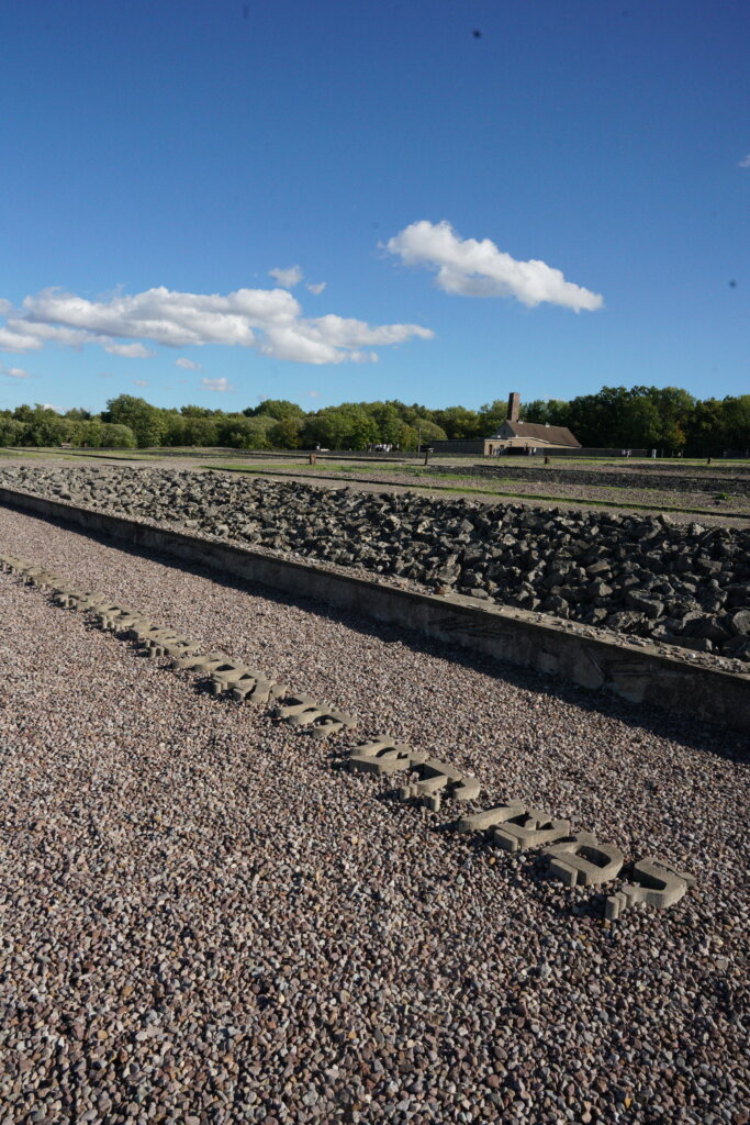 Stones of Hebrew letters lay in the ground in front of a destroyed bunk filled with debris. In the far background is a Holocaust-era crematorium.