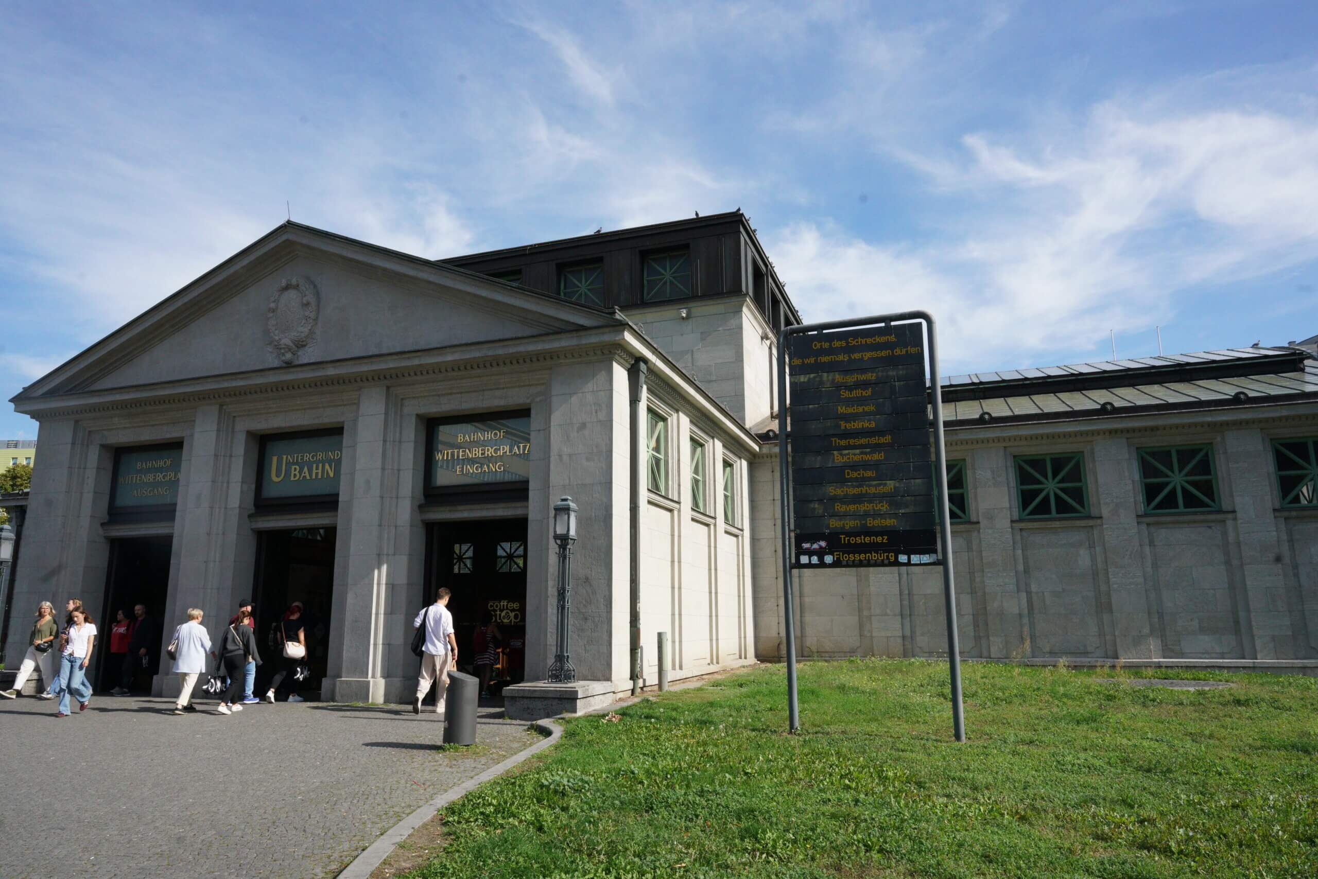 People walk into a large 19th century train station. On the right, a sign displays the names of Holocaust concentration camps