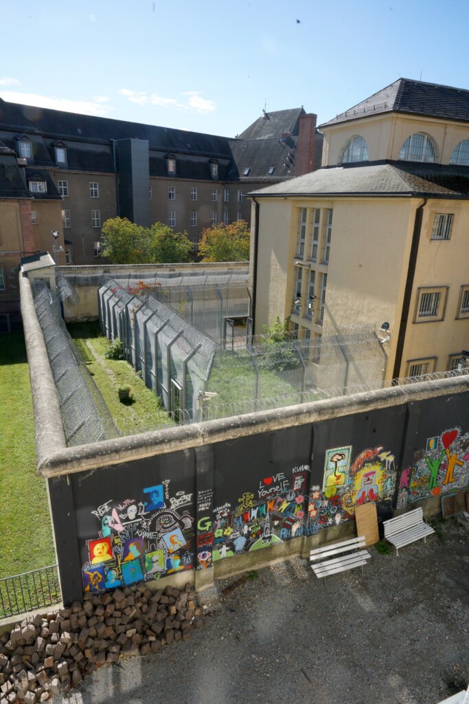 A prison sits behind a barbed wire fence and high exterior wall, which is covered in colorful paintings and graffiti on the outside.