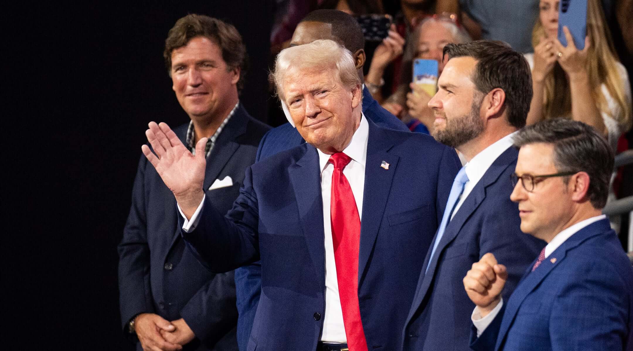 Former President Donald Trump waves to the crowd at the Republican National Convention at the Fiserv Forum in Milwaukee, Wisc., on Monday, July 15, 2024. Trump is flanked from left by Tucker Carlson, Rep. Byron Donalds, R-Fla., Vice-President nominee Sen. J.D. Vance, R-Ohio, and Speaker of the House Mike Johnson, R-La. 