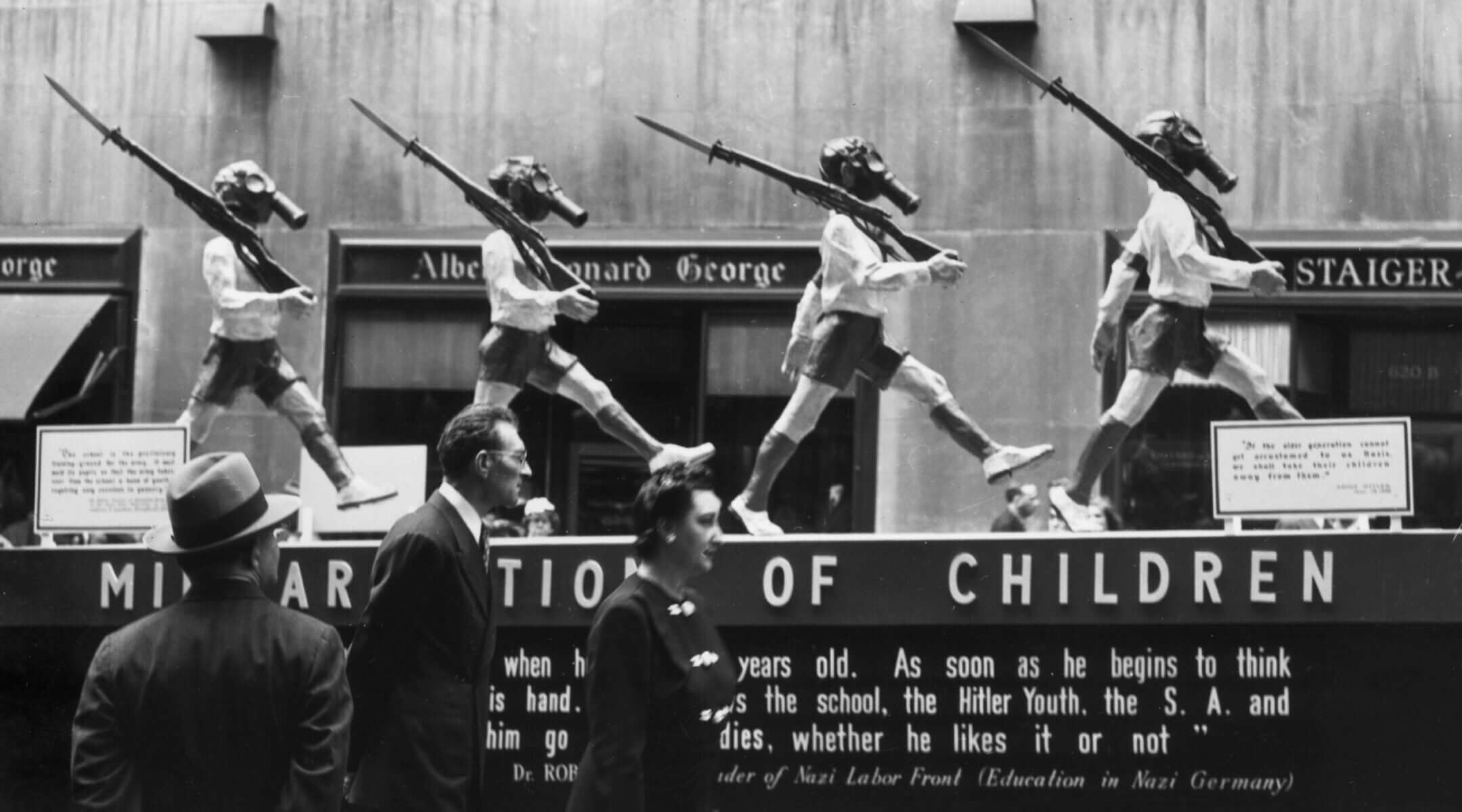 People view an anti-Nazi display with statues of young boys wearing shorts, armbands, gas masks and carrying rifles, Rockefeller Center, New York City. 