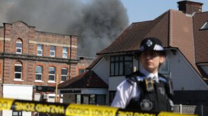 A member of the police stands at a cordoned area as smoke rises in the background from a fire that broke out at a kosher supermarket in the Golders Green neighbourhood in north London, May 27, 2026.